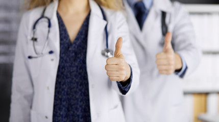 Two doctors standing with thumbs up in hospital office. Medical help, countering viral infection and medicine concept