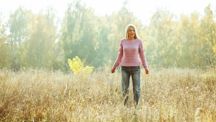 Charming blonde standing alone on the lawn among autumn or summer nature backlit by sunlight. Relaxation concept, Freedom concept