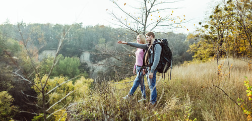 Tourists visiting surroundings from top of hill. Caucasian man with backpack points his hand into distance, charming blonde looks carefully. Traveling on foot concept