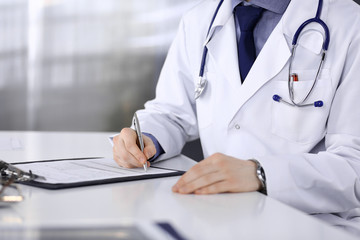 Unknown male doctor sitting and working with clipboard of medication history record in clinic at his working place, close-up. Young physician at work. Perfect medical service, medicine concept