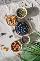 Top view of breakfast ingredients like cereal, almond, blueberry, pepitas and chia seeds on a linen cloth decorated with palm leaf.
