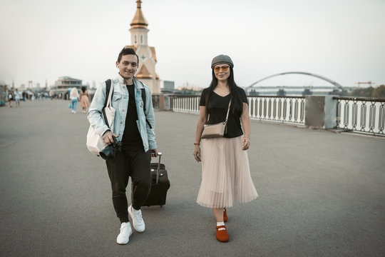 Two travelers are walking along the promenade of a european city. Smiling man and woman with backpacks and suitcases go along the riverbank looking at camera. Adventure concept