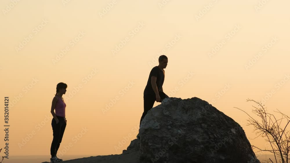Wall mural Man and woman hikers helping each other to climb stone at sunset in mountains. Couple climbing on high rock in evening nature. Tourism, traveling and healthy lifestyle concept. - Wall murals