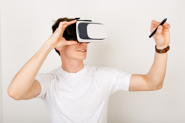 Male using virtual reality headset interacts with augmented things orienting in three dimensional space while sitting on his desk with keyboard and laptop on white background.