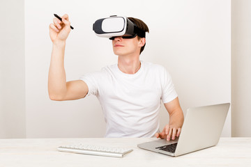 Male using virtual reality headset interacts with augmented things orienting in three dimensional space while sitting on his desk with keyboard and laptop on white background.