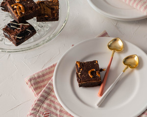 Top view down onto serving of delicious tempting brownie on a white plate with pink and white background.  Stacked brownies to right side on glass serving dish.  