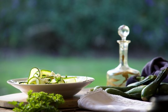 Zucchini ribbon salad on a wooden table .  An oil decanter and fresh whole zucchinis sit to the side of the bowl of salad.  
