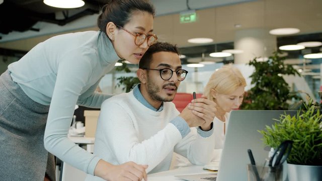 Male and female colleagues multi-ethnic team are talking looking at laptop indoors in shared office. Communication, coworking and people concept.