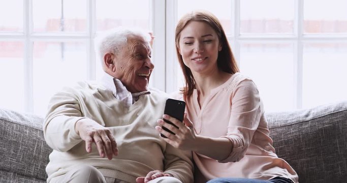 Happy older 80s grandfather posing for selfie shot on cellphone with smiling attractive grownup granddaughter, resting on sofa. Joyful different generations family having fun, using mobile photo apps.