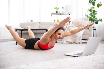 Fit woman doing superman's exercise and watching online workout tutorial on laptop, training in living room