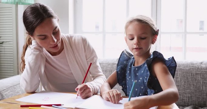 Head shot young mixed race female tutor teaching little girl drawing in paper album. Focused baby sitter nanny coloring pictures or preparing art lesson homework with school aged little girl.