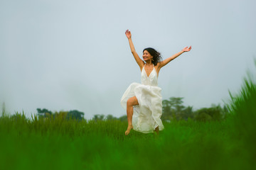 middle aged attractive and elegant Asian woman in white dress jumping happy and free enjoying beauty of nature at tropical green field playful and carefree