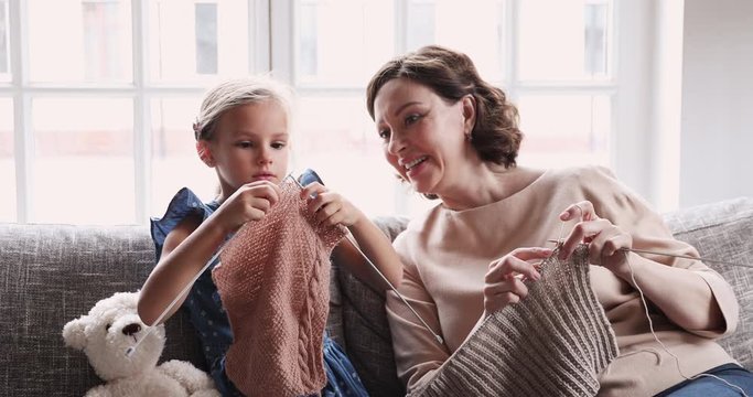 Attentive smiling middle aged woman teaching focused little granddaughter knitting warm scarf with wooden threads. Happy multigenerational family involved in hobby pastime, creating handmade clothes.