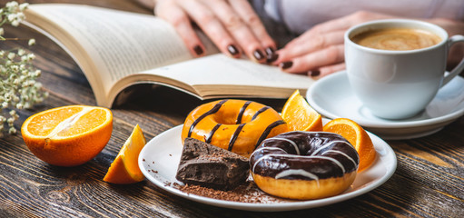 Woman is reading a book and having breakfast. Meal consists of cup of natural black coffee with fresh orange, donuts with orange and chocolate. Bright summer Breakfast, a tasty start to the morning