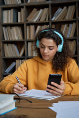 Hispanic teen girl school student wear headphones hold smartphone using distance learning mobile...