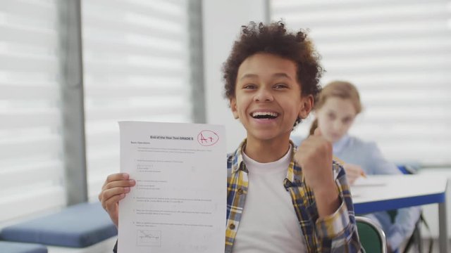 Lockdown of African school boy sitting at desk in classroom, holding sheet of paper and expressing joy because of excellent mark for test