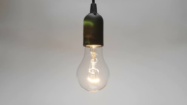 Classic glass lamp with tungsten filament in a black cartridge hangs on a black wire, lights up and flickers on a white background. Closeup. The camera moves around