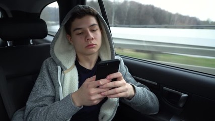 A young guy in a hood rides in the back seat of a car and is texting on the phone. Teenager and phone in a car with parents