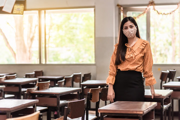 The Asian female teacher is wearing a mask and standing in the empty classroom because of living at risk in the midst of Coronavirus disease 2019 (COVID-19) epidemic.