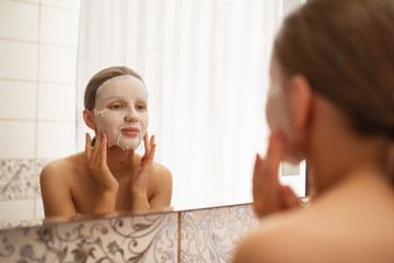 Obraz na płótnie Canvas A beautiful woman smoothes, puts on a face a cosmetic mask in the bathroom by the mirror. Healthy facial skin. Pore cleansing. Skin care. Mask sheet