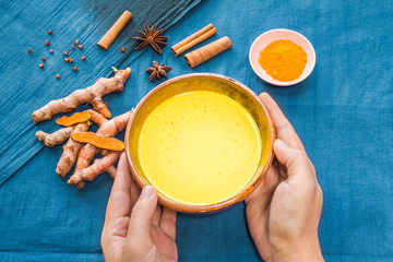 Hands holding organic curcumin honey golden milk, Indian turmeric latte with ingredients on bright blue background. Top view exotic spices yellow natural drink healthy food concept