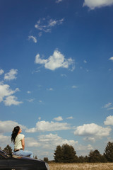 Young woman traveler sitting on the car hood while traveling. Road trip, vacation and adventures