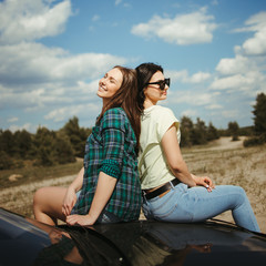 Traveling, camping, open air party. Young women friends traveler sitting on the car hood while traveling. Road trip,friendship, vacation and adventures