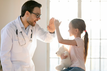 Smiling little girl patient give high five have fun at consultation with excited male doctor or pediatrician, happy small child client greeting with man GP in hospital or clinic, healthcare concept