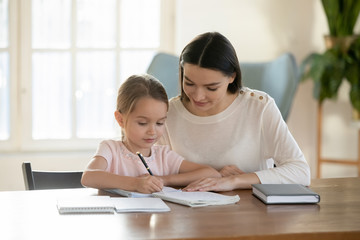 Caring young Caucasian mother sit at table with little daughter learn handwriting, loving mom or nanny help small girl child with school preparation, studying teaching at home, education concept