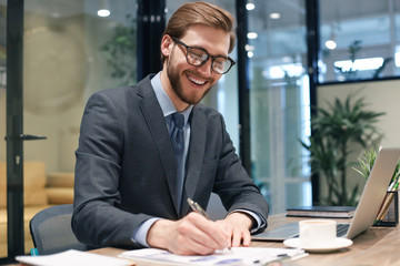 Smiling young business man writing something down while working in modern office.