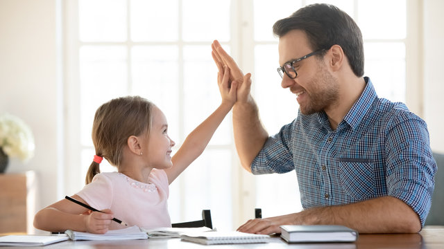 Overjoyed dad and little preschooler daughter give high five celebrate success in studying, smiling father greeting cheering small girl child writing or learning at home together, education concept
