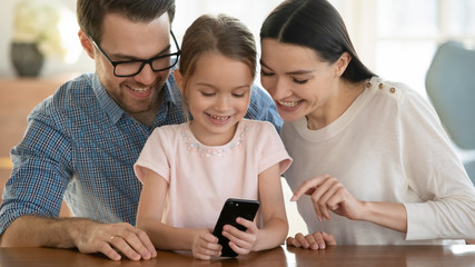 Happy young family with little daughter sit at table browsing wireless internet on modern smartphone together, smiling parents with small girl child have fun using cellphone gadget at home