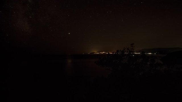Top view of landscape of bay of mountains over Black Sea with luminous houses and lanterns under bright shining moon and moving starry sky at night in Crimea, time lapse of coastal moonlight path