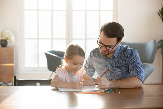 Loving young father sit at desk studying drawing with small preschooler daughter, caring happy dad have fun painting picture with little girl child, enjoy leisure family weekend at home together