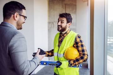 Handsome Caucasian bearded construction worker in work wear talking with his boss about project. Building in construction process interior.