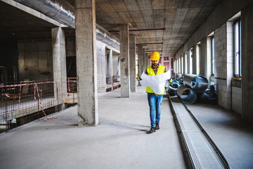 Full length of construction worker in work wear walking in building in construction process and looking at blueprints.a