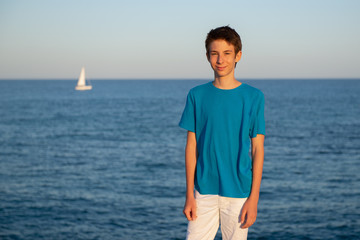 Handsome young happy boy at beach. Beautiful calm smiling teen boy posing alone at Mediterranean sea coast with sailboat at background. Travel, summer vacation, tourism, teenage lifestyle.