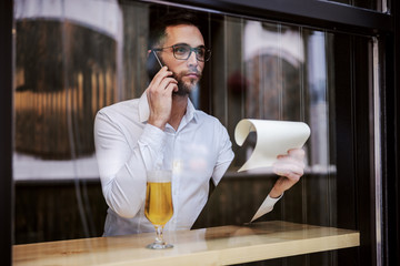 Young serious businessman in shirt sitting in pub after work, holding paperwork and calling boss after work.