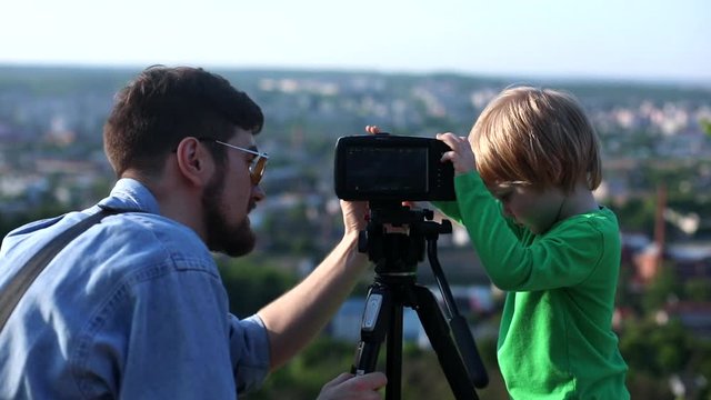 Young stylish father with cute little son enjoying learning to use professional camera together with city landscape on sunny background. Freelance business lifestyle. Father Day. Early kids education.
