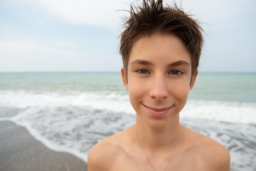 Handsome young boy at beach, summer vacation, wide angle. Funny portrait of beautiful smiling teen boy at sea coast posing after swimming. Travel, summer vacation, tourism, recreation