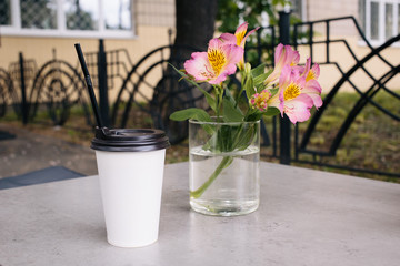 A white paper cup with lid and black straw on the table in the summer terrace cafe. Latte coffee or tea. Eco. break. Summer day. A flowers on the glass.