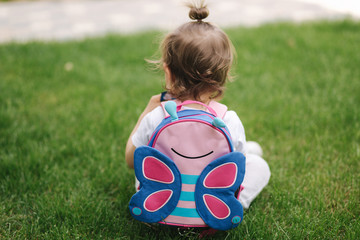 Back view of little girl with backpack sits on the grass outdoors. space for text