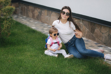 Mom with her adorable little daughter sits on the grass in front of house. Happy family spent time outdoors. Mother and daughter in sunglasses