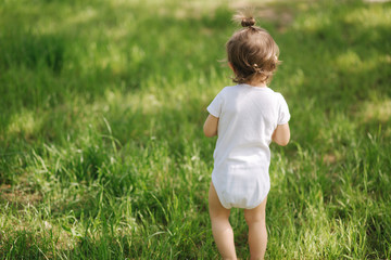 Back view of Adorable little girl in white bodysuit walk in backyard on the grass barefoot