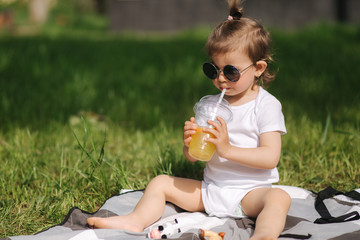 Happy little kid sits outdoors on carpet and drink lemonade. Adorable little girl in white bodysuit