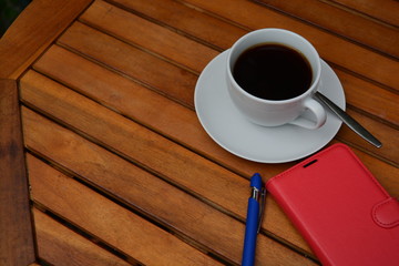 a cup of black coffee in a white cup in the morning on wooden table with  red cover telephone and blue pen