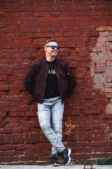 Young man in jeans, sneakers and jacket standing with his hands in his pockets by a brick wall.