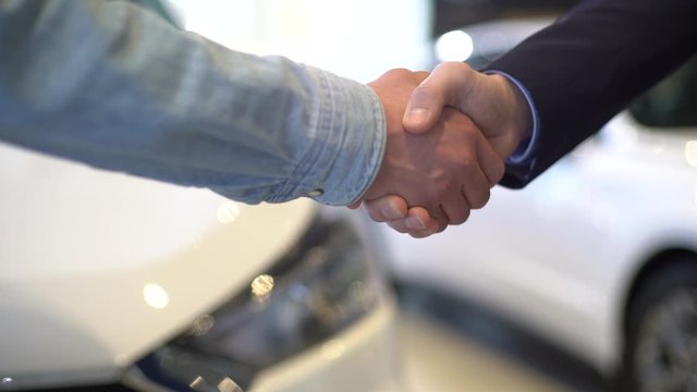 Close-up of unrecognizable salesman giving car keys to customer and shaking his hand in auto dealership, in front of car. Concept of buying new car at showroom. Shooting in slow motion.