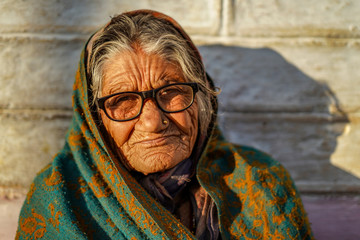 Old aged woman sitting with the support of the wall in the sunset wearing glasses