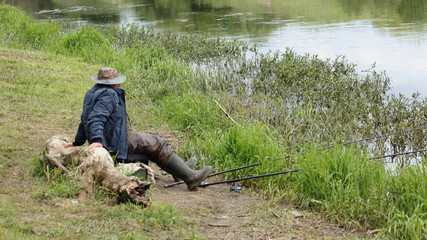Elderly fisherman in camouflage hat clothing with two fishing rod sitting in green grass on the river Bank on a spring day, calm outdoor fishing recreation, side view